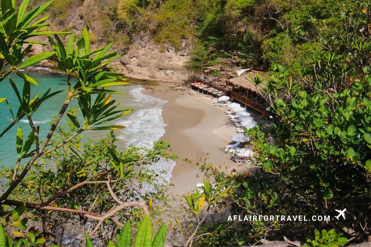 Smuggler’s Cove beach at Cap Maison in St. Lucia with white sand, turquoise water, and lounge chairs beneath a cliffside restaurant.