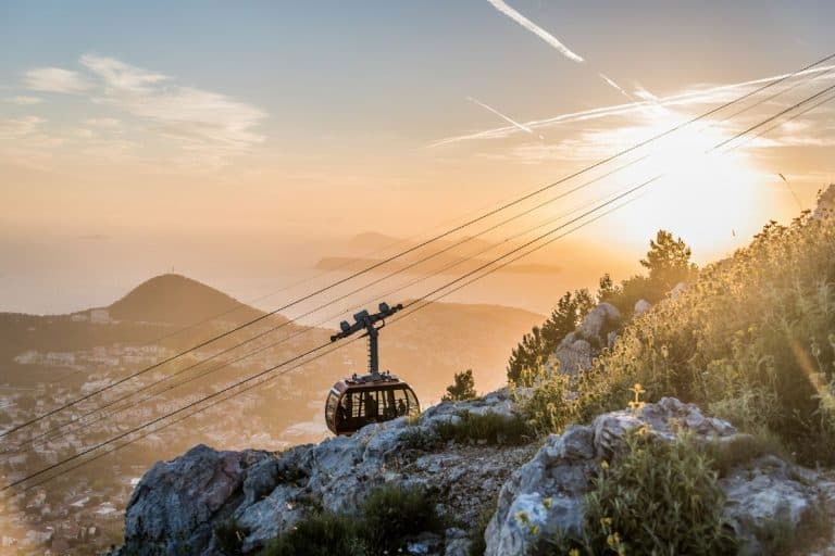 Cable car ascending Mount Srđ at golden hour with panoramic views of Dubrovnik coastline, mountains, and sunset sky in background; a popular thing to do in Dubrovnik is to watch the sunset from the top of Mt. Srd.