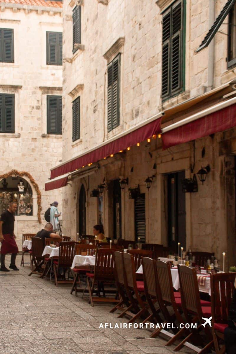 Charming cobblestone alleyway in Dubrovnik with outdoor cafe tables, red awning with string lights, green shutters, and historic stone architecture; one of the things to know about Dubrovnik is that advance reservations are needed since many restaurants are so small.