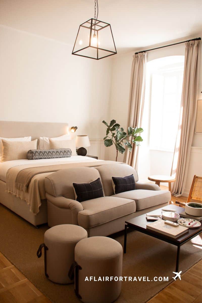 Modern hotel room interior in Dubrovnik with neutral tones, upholstered bed, beige sofa, geometric pendant light, round ottomans, and bright windows with linen curtains; one of the things to know about Dubrovnik is that some hotels don't have elevators.