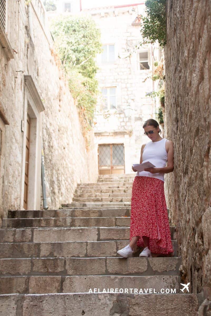 Woman reading map on stone stairs in historic Dubrovnik alley between tall limestone walls with potted plants and traditional architecture