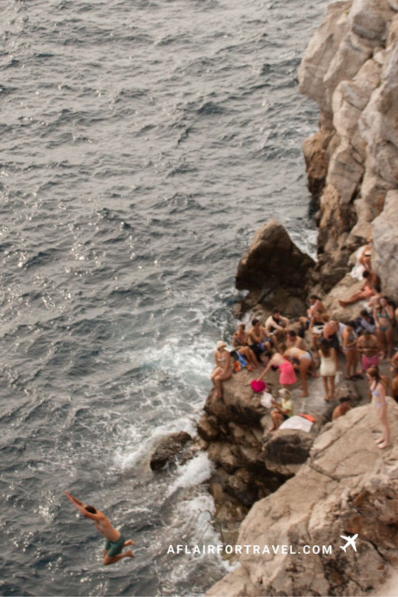 Cliff diving spot in Dubrovnik with swimmers on rocky ledges and person jumping into the Adriatic Sea; one of the things to know about Dubrovnik is that this can be dangerous.
