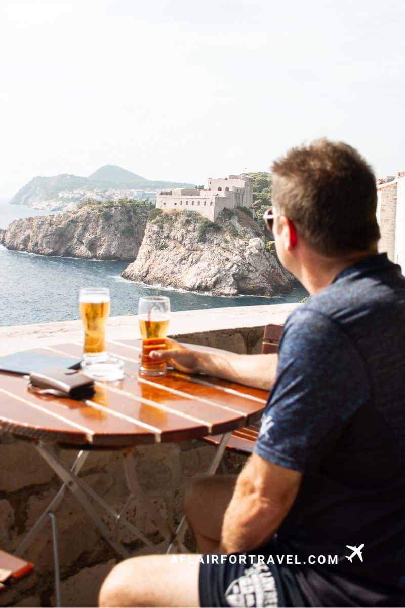 Man enjoying beer at outdoor cafe table on the walls, with panoramic view of Lovrijenac Fortress perched on cliffs above the Adriatic Sea in Dubrovnik. Visitors need to know how hot it can get.