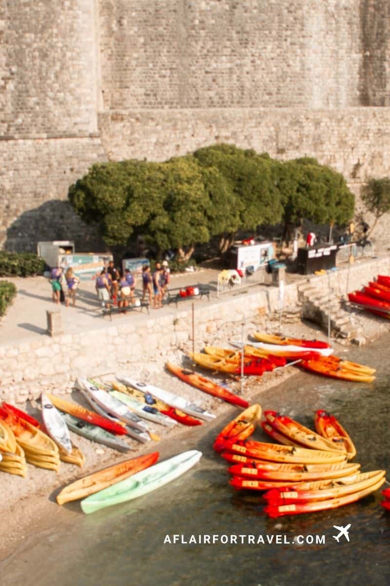 Colorful kayaks lined up on small beach beneath medieval stone fortification walls in Dubrovnik, with tourists and rental equipment visible