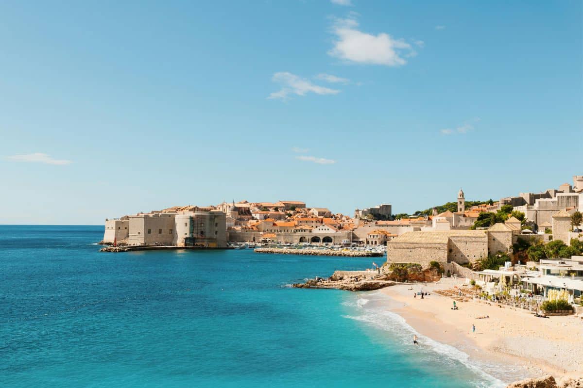 Banje Beach in Dubrovnik with turquoise Adriatic waters, sandy shoreline, beachgoers, and medieval walled Old Town with stone fortifications rising from the sea under blue sky