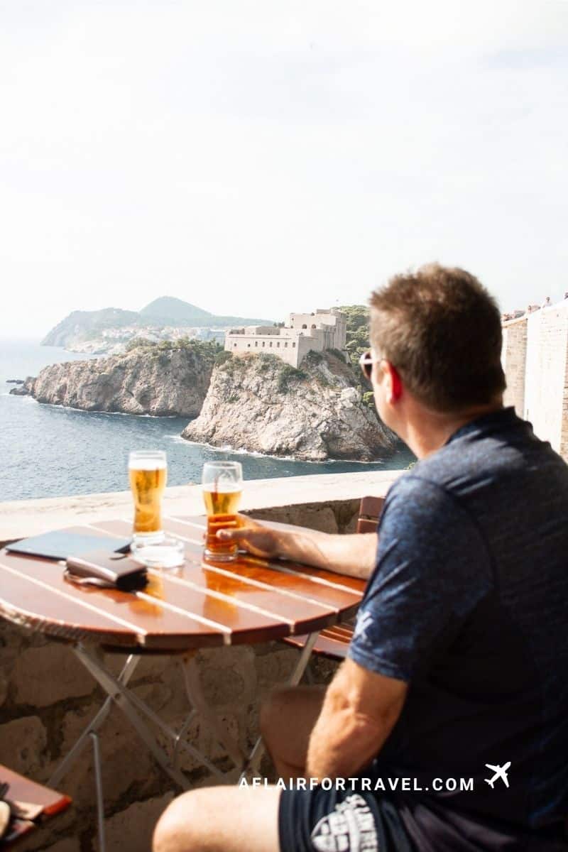 Man enjoying a cold beer at an outdoor bar on Dubrovnik's ancient city walls with views of Fort Lovrijenac and the Adriatic Sea