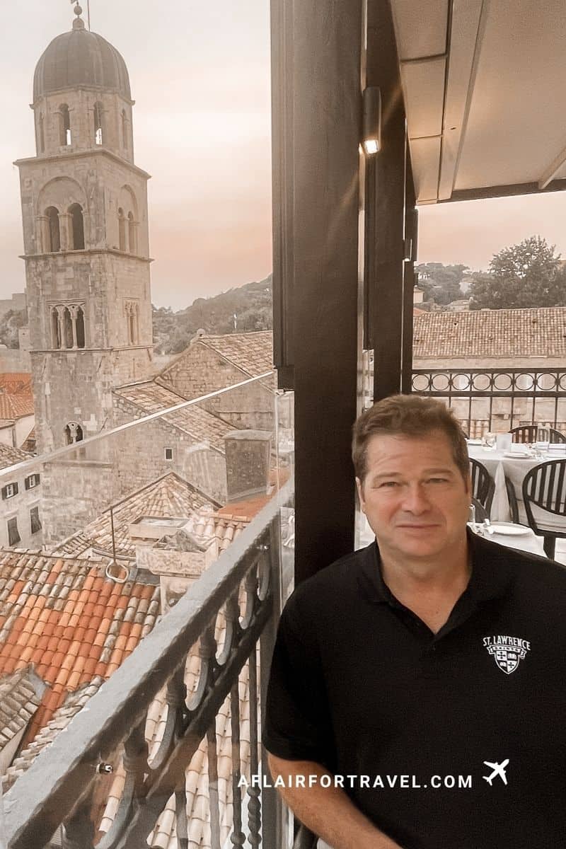 Man sitting next to a restaurant balcony overlooking Dubrovnik's historic rooftops and bell tower at sunset
