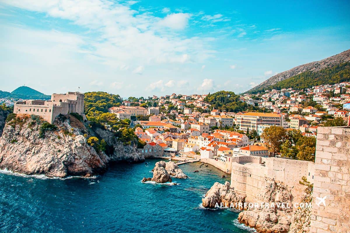 Panoramic view of Dubrovnik's Old Town from the city walls, showing the historic stone fortifications, terracotta-roofed buildings, and the deep blue Adriatic Sea, with forested hills in the background.