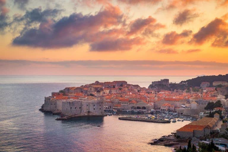 Panoramic sunset view of Dubrovnik's Old Town with glowing orange rooftops, ancient stone walls, and a harbor full of boats along the Adriatic Sea