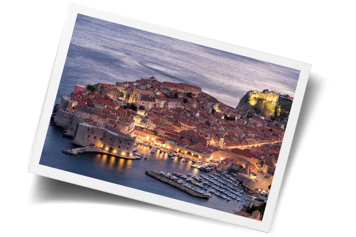 Elevated view of Dubrovnik Old Town at dusk, showing the illuminated harbor, terracotta rooftops, and Fort Lovrijenac on the cliffs