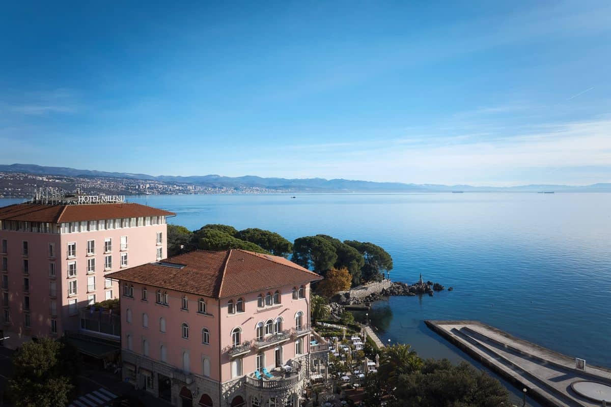 Aerial view of Amadria Park Hotel Milenij, a pink heritage building set on the Adriatic coastline in Opatija, Croatia, with calm blue sea and mountains in the background.