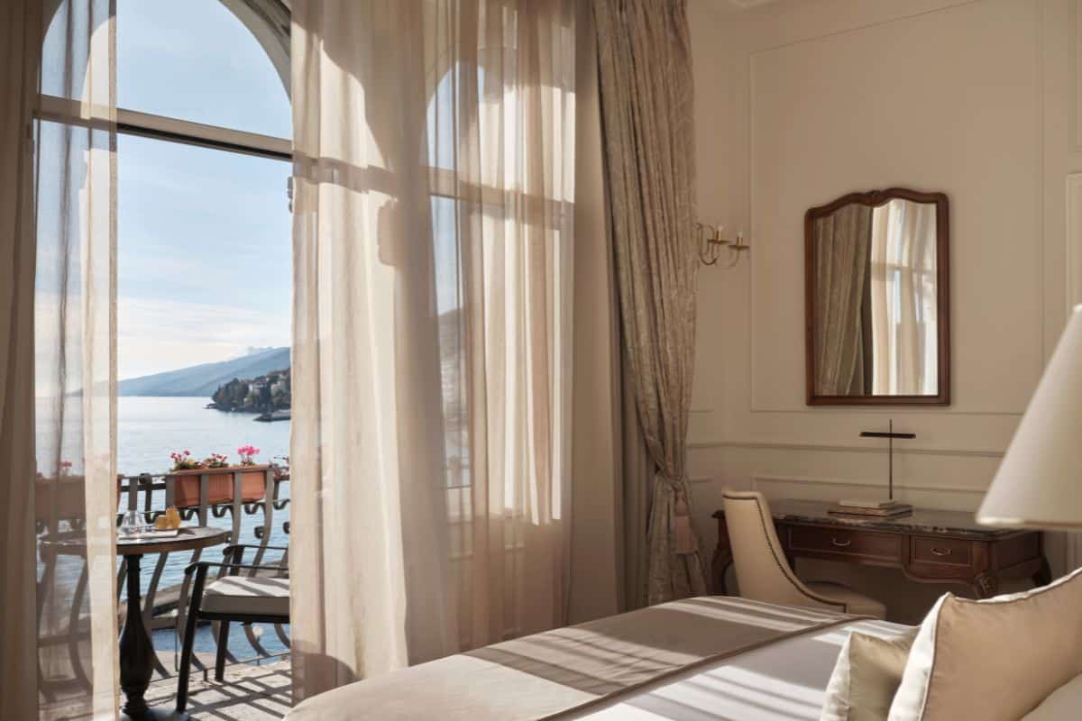 A elegantly decorated hotel room at Amadria Park Hotel Milenij, featuring sheer linen curtains, a classic wooden desk and mirror, and an arched balcony door opening to a sea view.