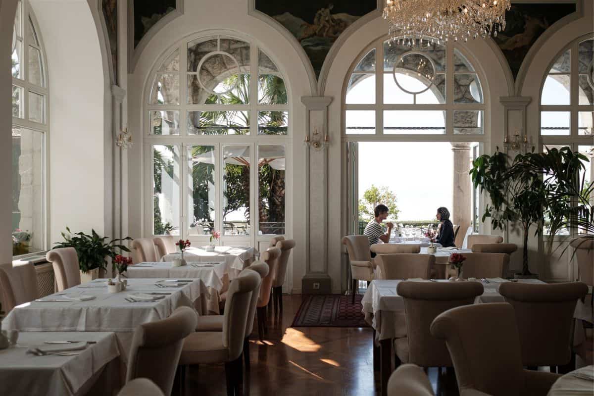 The grand restaurant interior at Amadria Park Hotel Milenij, with ornate arched windows, painted ceilings, crystal chandelier, and white-clothed tables overlooking the Adriatic Sea.