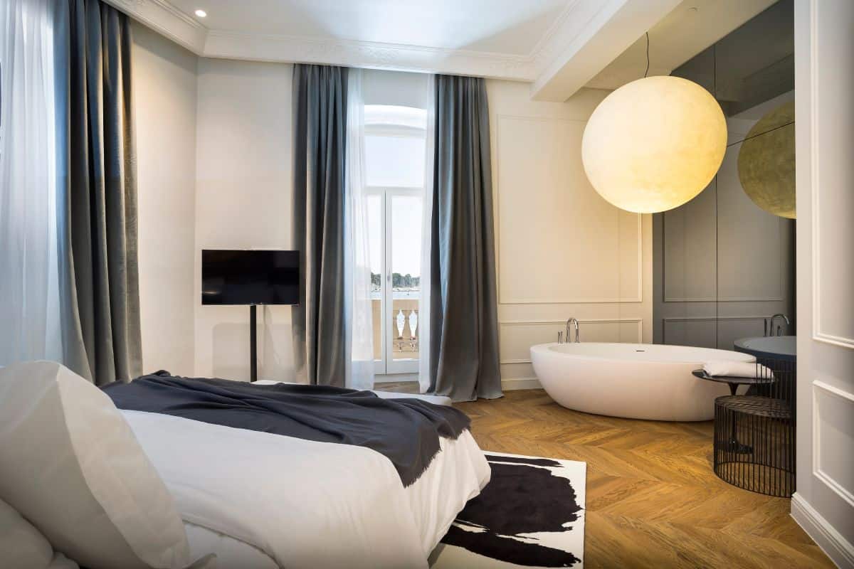 A stylish suite at Adriatic Hotel Rovinj with a king bed, herringbone parquet floor, floor-length grey curtains, a freestanding bathtub, and a large globe pendant light, with a balcony overlooking the harbour.