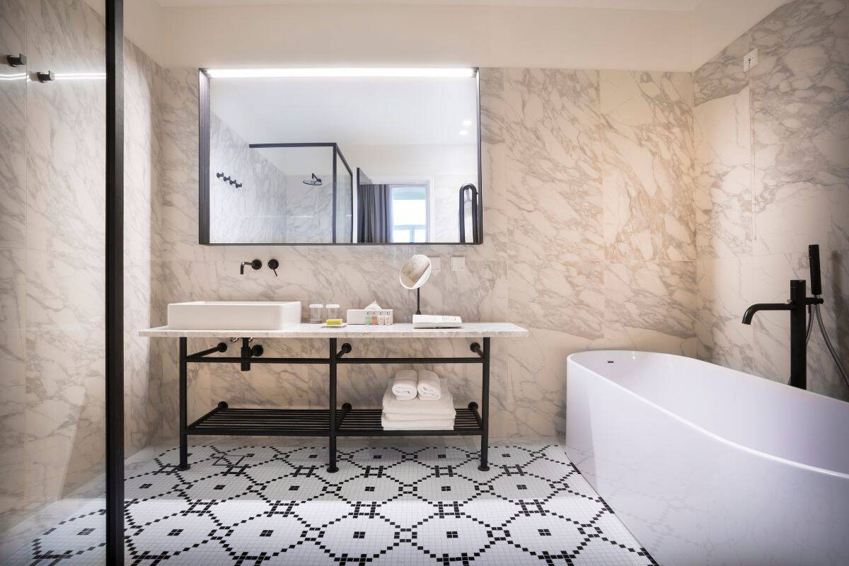 A luxury bathroom at Adriatic Hotel Rovinj featuring marble-clad walls, a black and white mosaic tile floor, an open vanity unit with black metal frame, and a freestanding bathtub with a black floor-mounted tap.