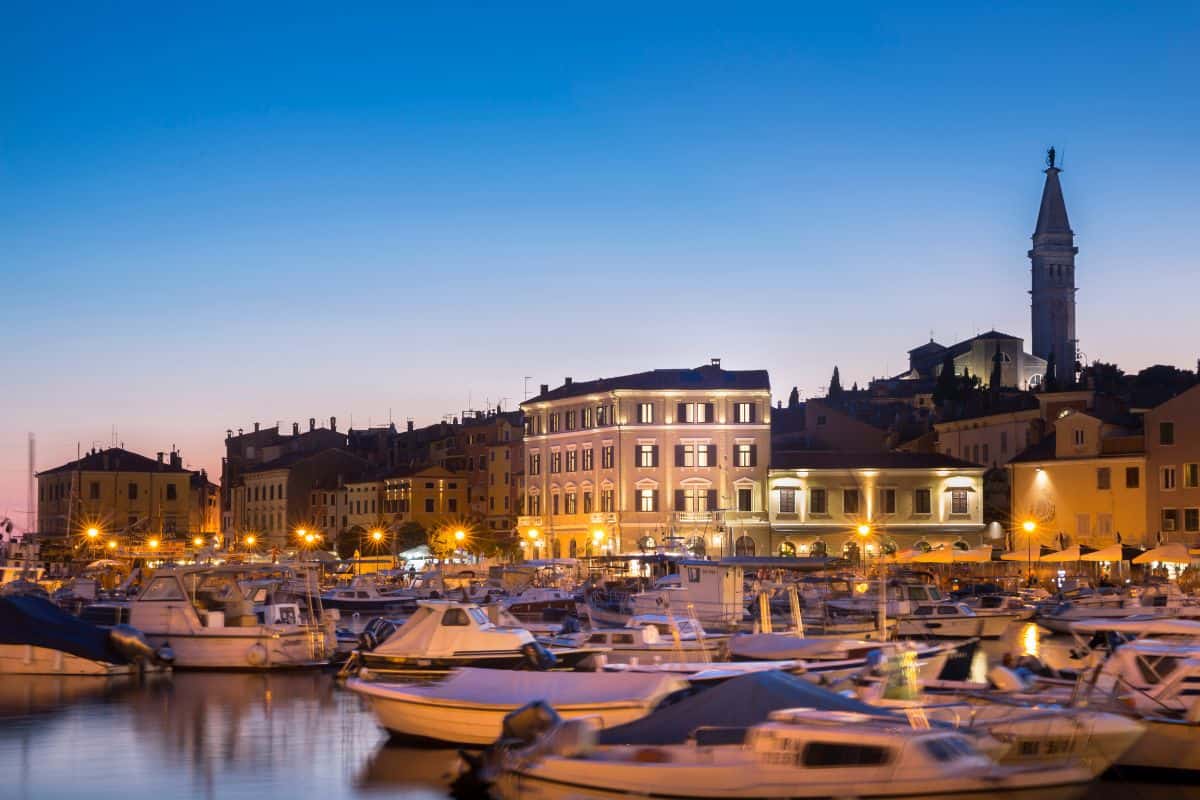 The illuminated facade of Adriatic Hotel Rovinj, a boutique hotel in Croatia, seen from the harbour at dusk, with boats moored in the foreground and the bell tower of the Church of St. Euphemia rising above the old town rooftops.