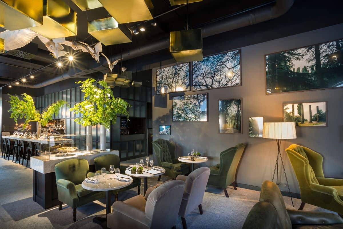 The dramatic restaurant interior at Adriatic Hotel Rovinj, featuring dark walls, a gold geometric ceiling, lush indoor trees, sculptural white birds in flight, velvet armchairs in shades of green, and an open kitchen counter.