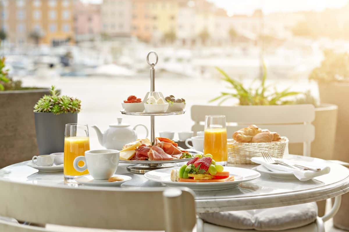 A breakfast spread laid out on a harbourside terrace table at Adriatic Hotel Rovinj, with fresh fruit, prosciutto, pastries, orange juice and tea, with the marina visible in the soft morning light behind.