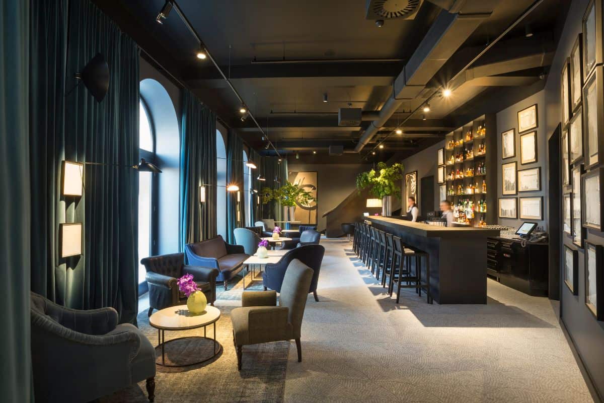 The moody bar and lounge area at Adriatic Hotel Rovinj, with tall arched windows dressed in teal curtains, velvet armchairs, a long dark bar lined with spirits, and framed artwork covering the walls.