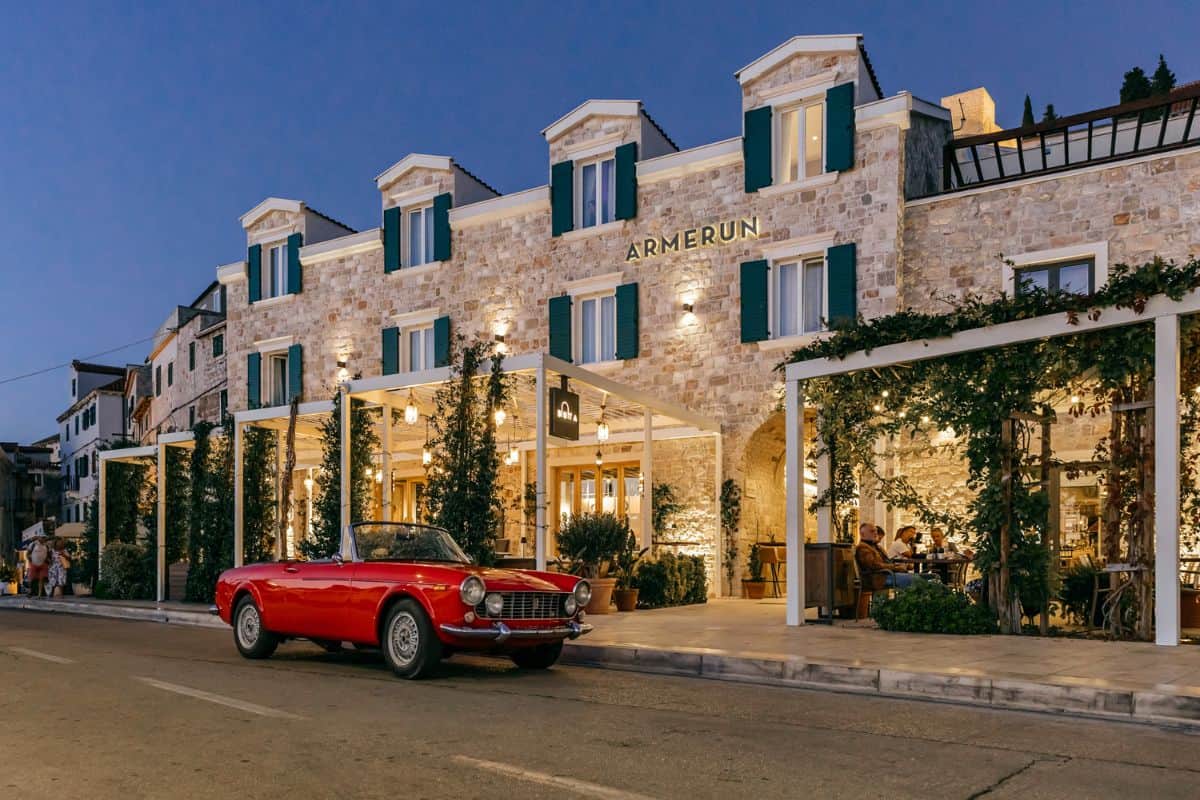 The illuminated stone facade of Armerun Hotel, a boutique hotel in Croatia, at dusk, with a red vintage convertible parked outside, climbing plants framing the entrance, and guests dining on the terrace.