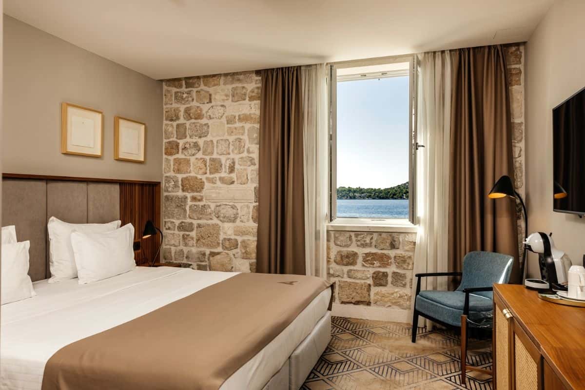 A hotel room at Armerun Hotel with a king bed, exposed stone wall, patterned tile floor, and an open window framing a direct sea view.
