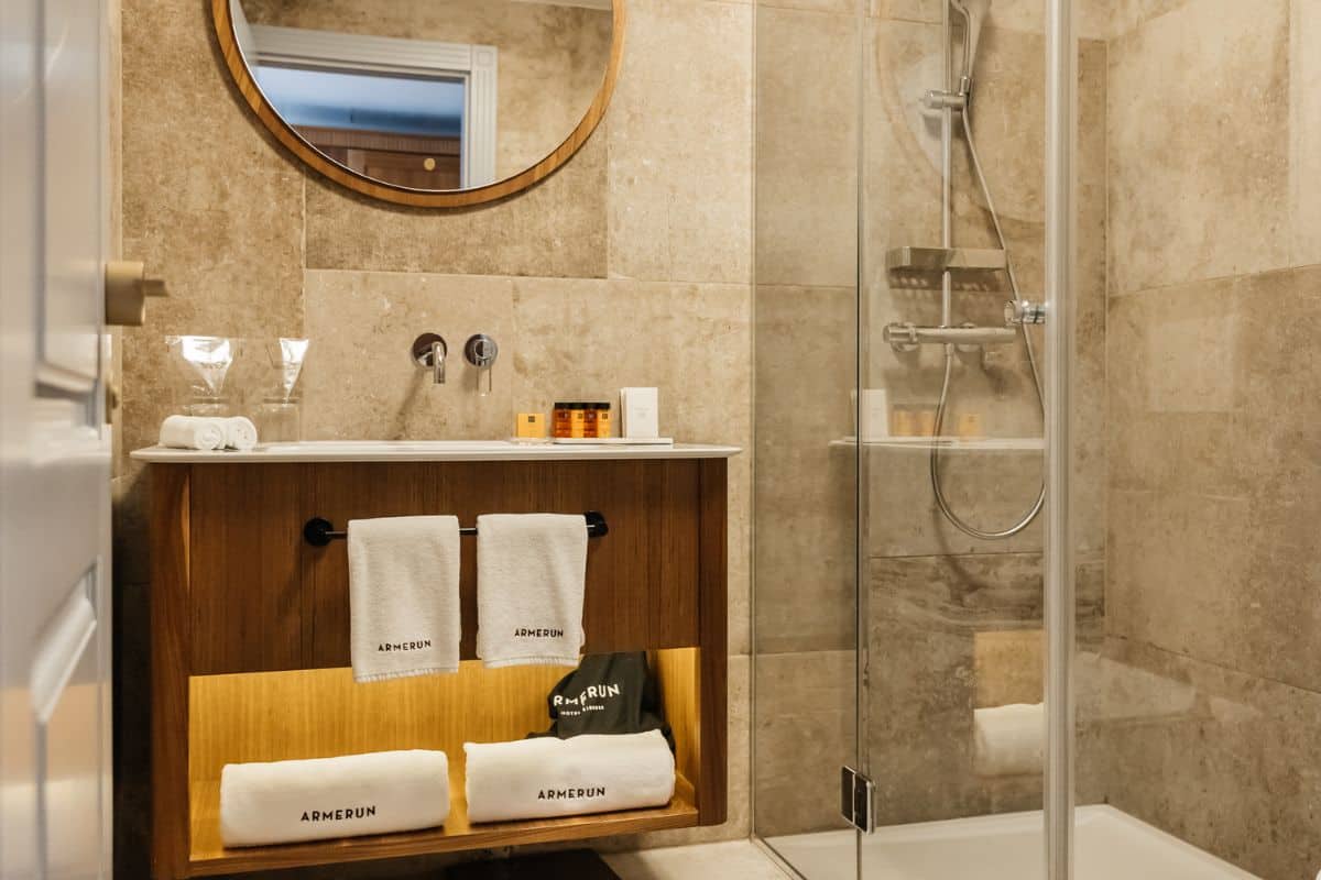 A bathroom at Armerun Hotel featuring travertine stone tiles, a wooden vanity unit stocked with Armerun-branded towels and toiletries, a round mirror, and a glass-enclosed shower.