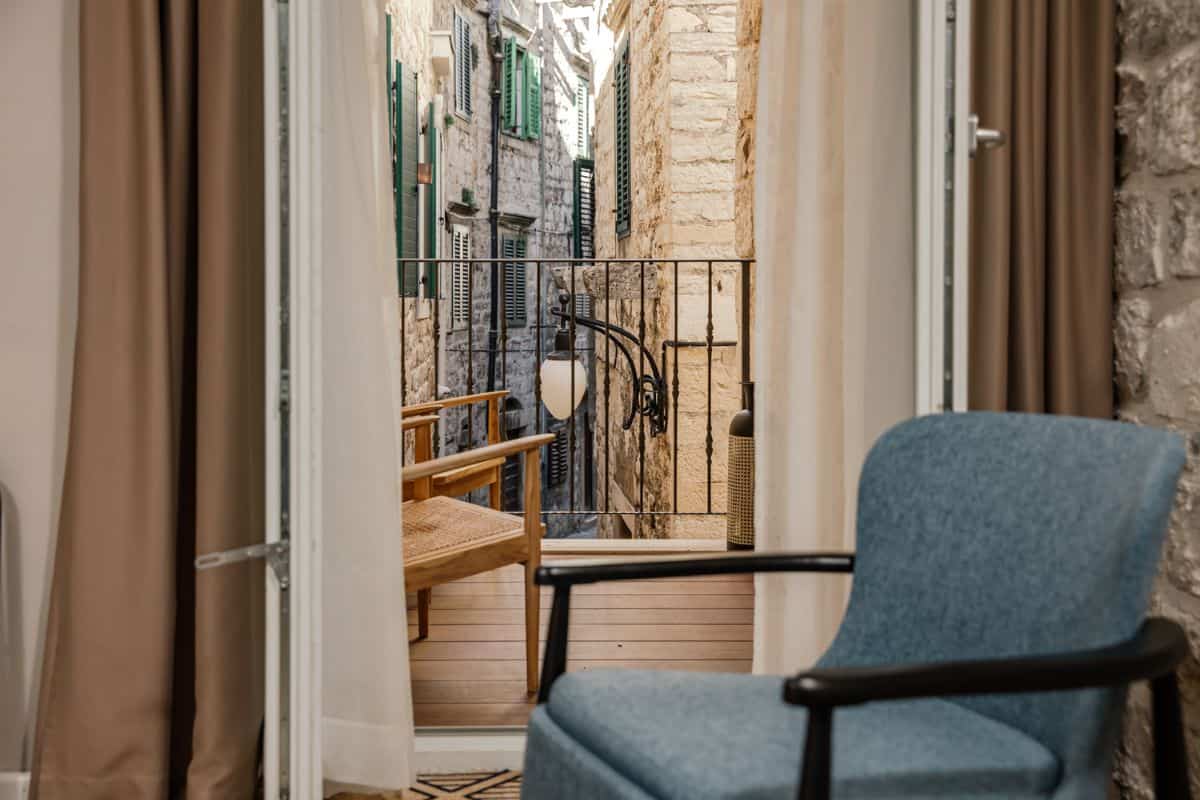 View from inside an Armerun Hotel room through open French doors to a small balcony overlooking a narrow stone-walled alley in the old town, with green shuttered buildings on either side.