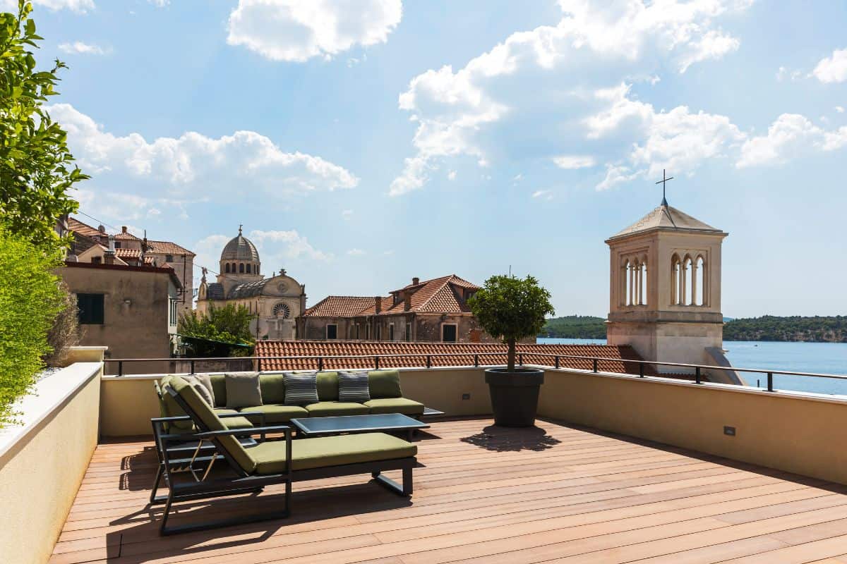 The rooftop terrace at Armerun Hotel with sun loungers and a green sofa set, looking out over the terracotta rooftops of Šibenik, the dome of the Cathedral of St. James, and the Adriatic Sea beyond.