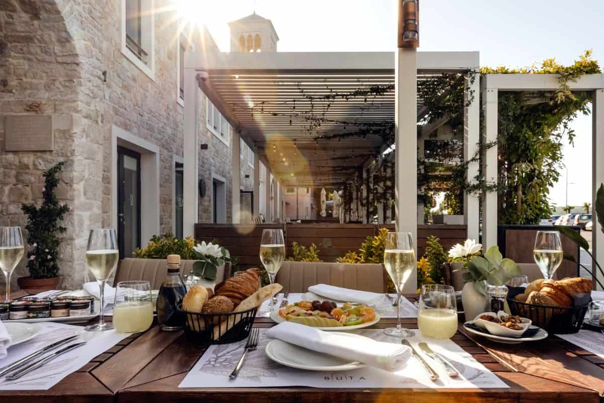 A sun-drenched outdoor breakfast table at Armerun Hotel's Buita restaurant, set with champagne flutes, fresh bread, fruit, and pastries, with a vine-covered pergola terrace stretching behind.