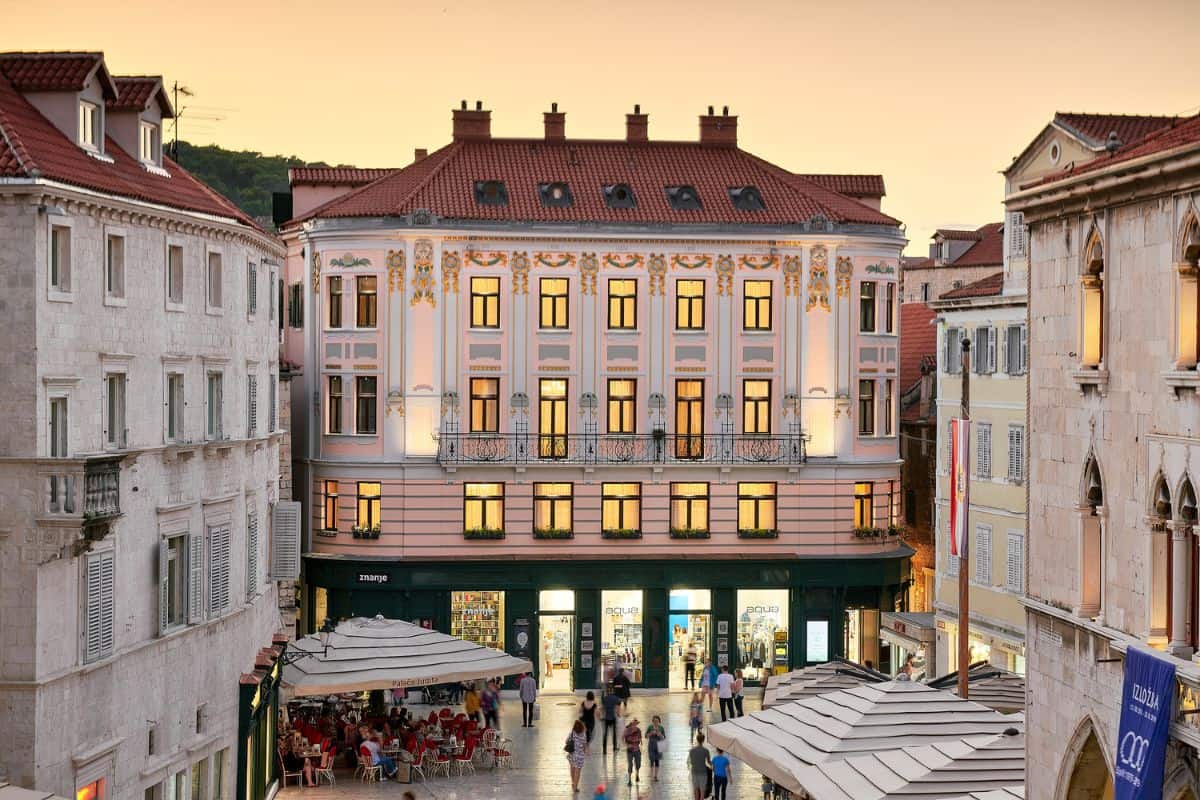 The ornate Art Nouveau facade of Piazza Heritage Hotel, a boutique hotel in Croatia, illuminated at sunset, overlooking a busy pedestrian square in the heart of Split's old town.