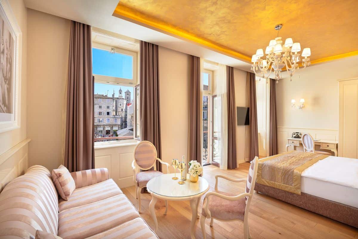 A spacious suite at Piazza Heritage Hotel Split with a gold leaf ceiling, Murano glass chandelier, striped sofa, circular dining table, and large windows overlooking the old town rooftops and clock tower.