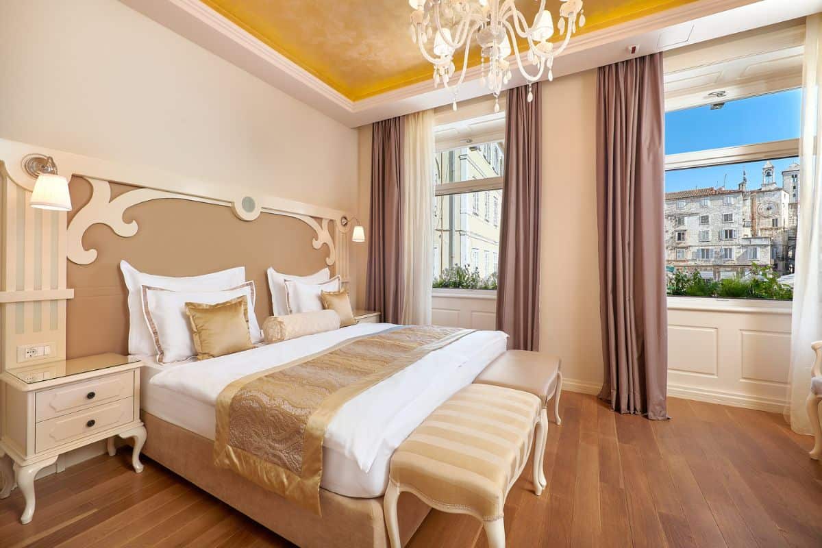 An elegant bedroom at Piazza Heritage Hotel Split featuring a decorative carved headboard, gold satin bedding, a Murano glass chandelier, and a large window with views over Split's old town.