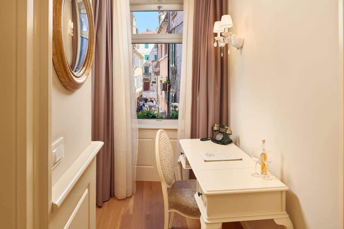A classic writing desk area in a room at Piazza Heritage Hotel Split, with a vintage rotary telephone, wine and glasses, a gilt oval mirror, and a window looking down onto a busy old town alleyway below