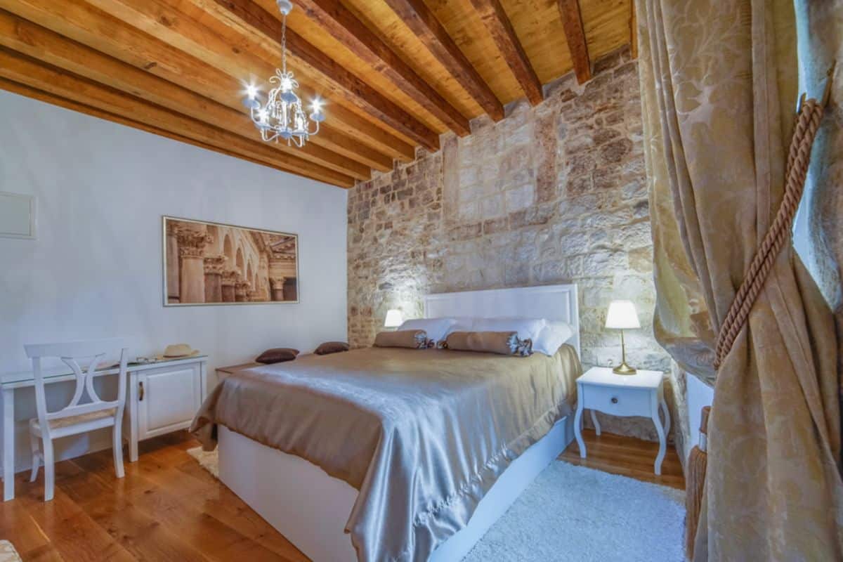 A hotel room at Villa Split Heritage Hotel, a boutique hotel in Croatia, with a double bed, silver satin bedding, exposed stone wall, wooden beam ceiling, small chandelier, and white painted furniture.