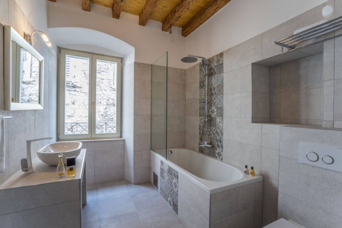 A bathroom at Villa Split Heritage Hotel with grey stone tiles, a vessel sink vanity, a bathtub with overhead rain shower, patterned tile detailing, and a wooden beam ceiling with an arched window.