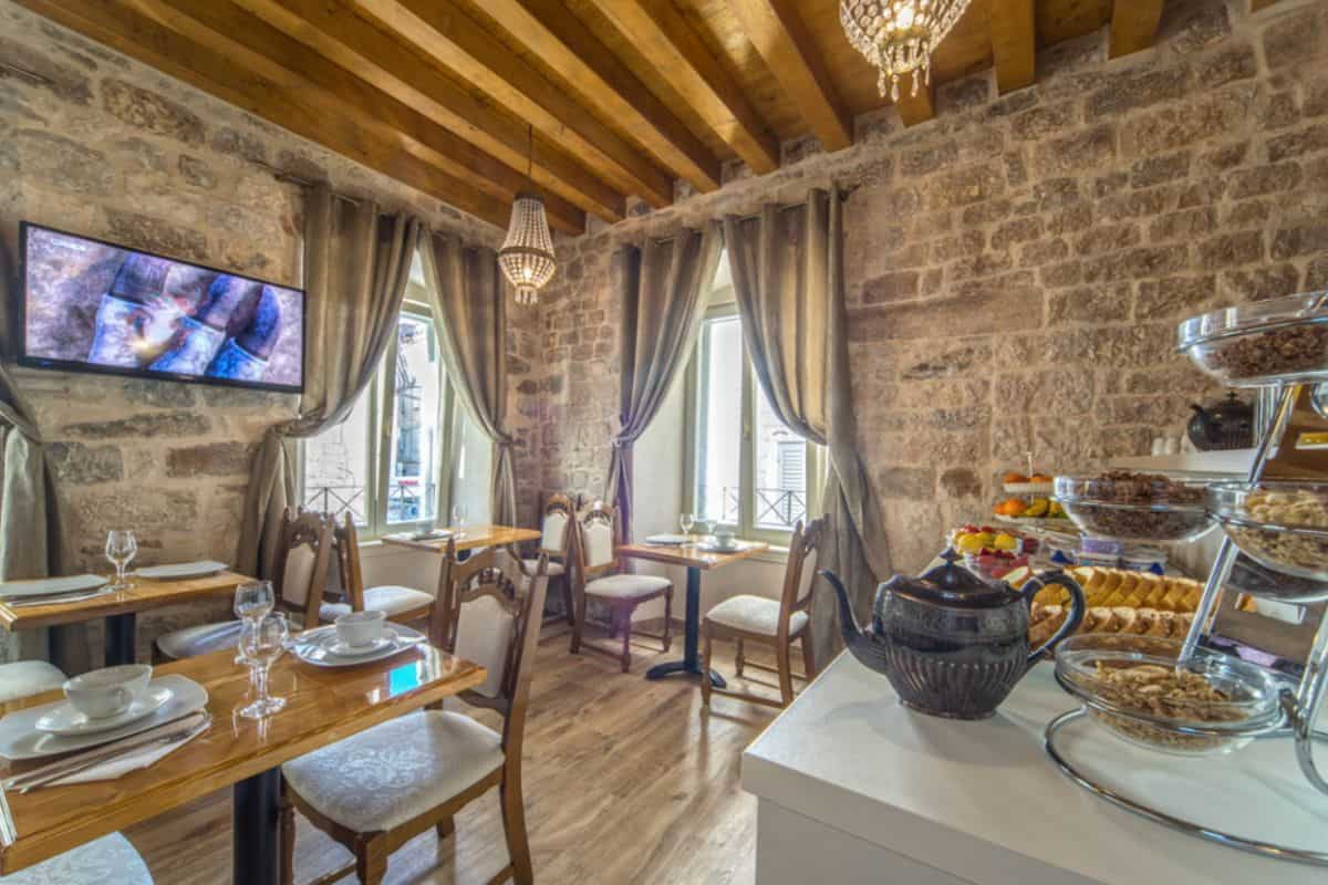 The breakfast room at Villa Split Heritage Hotel, a boutique hotel in Croatia, with exposed stone walls, wooden beam ceiling, crystal chandelier, set dining tables, and a buffet display of pastries, fruit and cereals.