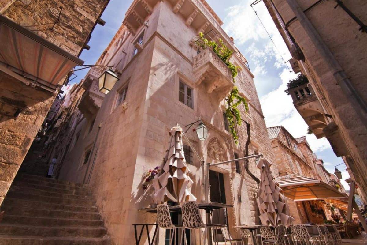 The historic stone exterior of Prijeko Palace, a boutique hotel in Croatia, viewed from a narrow alleyway in Dubrovnik's old town, with ornate carved detailing, a vine-draped balcony, and sculptural terrace umbrellas at street level.