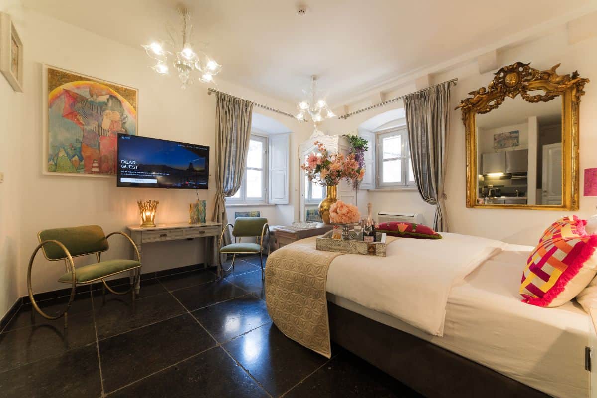A richly decorated room at Prijeko Palace Dubrovnik with a king bed, colourful original artwork, a large gilt mirror, Murano glass chandelier, green velvet chairs, and a welcome tray of champagne and flowers.