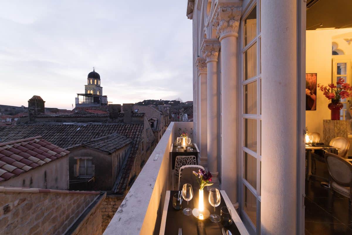 A candlelit balcony table set for dinner at Prijeko Palace Dubrovnik at dusk, with wine glasses and fresh flowers, ornate stone columns, and views across the old town rooftops to the illuminated cathedral dome
