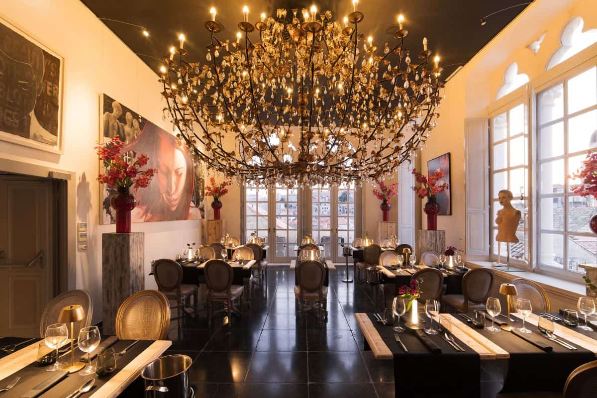The grand restaurant at Prijeko Palace Dubrovnik featuring a spectacular oversized chandelier, black tile floors, large-scale artworks, red floral arrangements, and tall windows overlooking the old town.