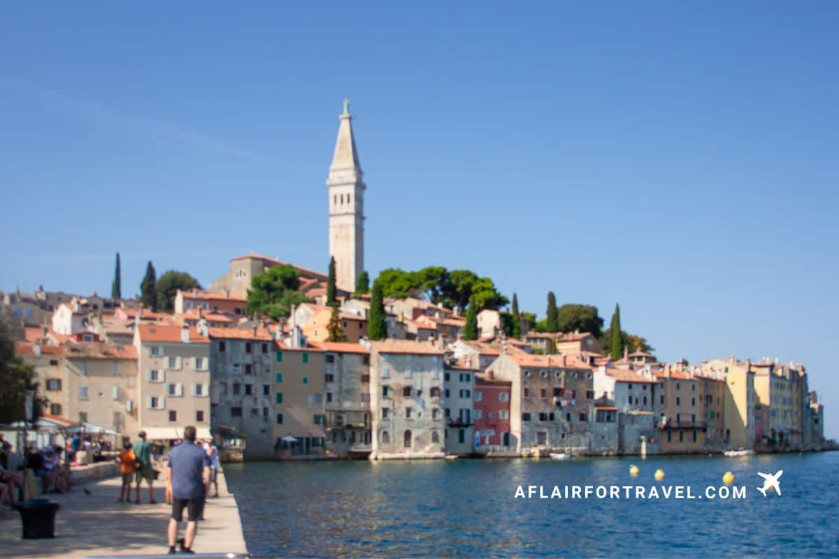 Rovinj waterfront lined with colorful historic buildings, a tall church bell tower, and people walking along the waterfront promenade on a sunny day in Croatia.