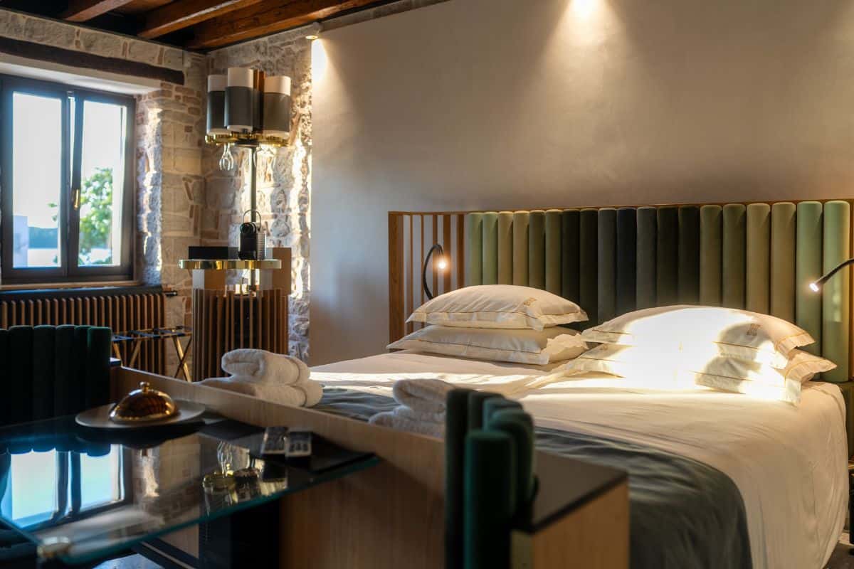 A boutique hotel room at Hotel Spirito Santo Palazzo Storico, a boutiqe hotel in Croatia, featuring a king bed with a tufted green velvet headboard, exposed stone walls, wooden beam ceiling, and warm accent lighting.