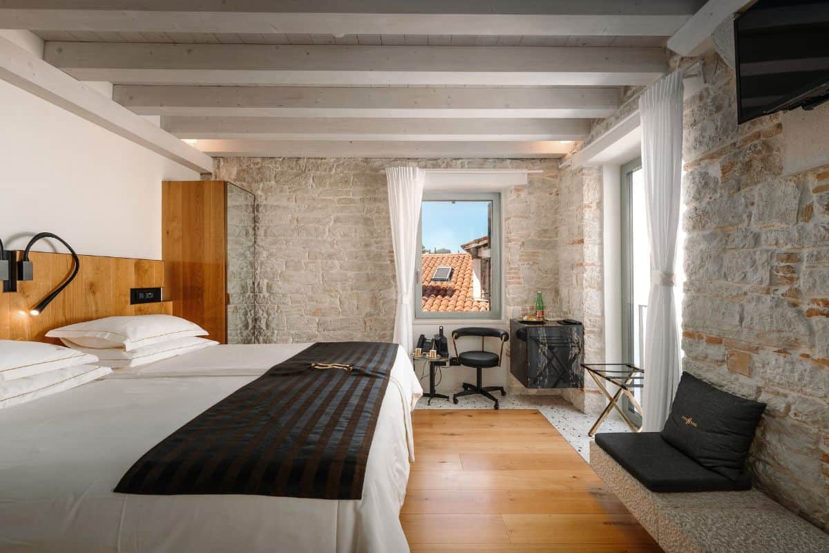 A bright hotel room at Hotel Spirito Santo Palazzo Storico with a wooden headboard, white bedding, exposed stone walls, light grey ceiling beams, and a window overlooking the old town rooftops.
