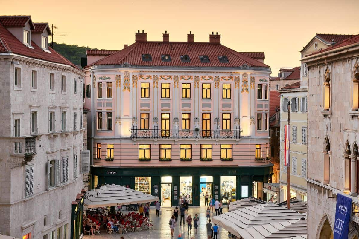 Historic square in Split, Croatia at sunset featuring Piazza Heritage Hotel's elegant pastel-colored building with illuminated windows, surrounding stone architecture, and outdoor café seating below.
