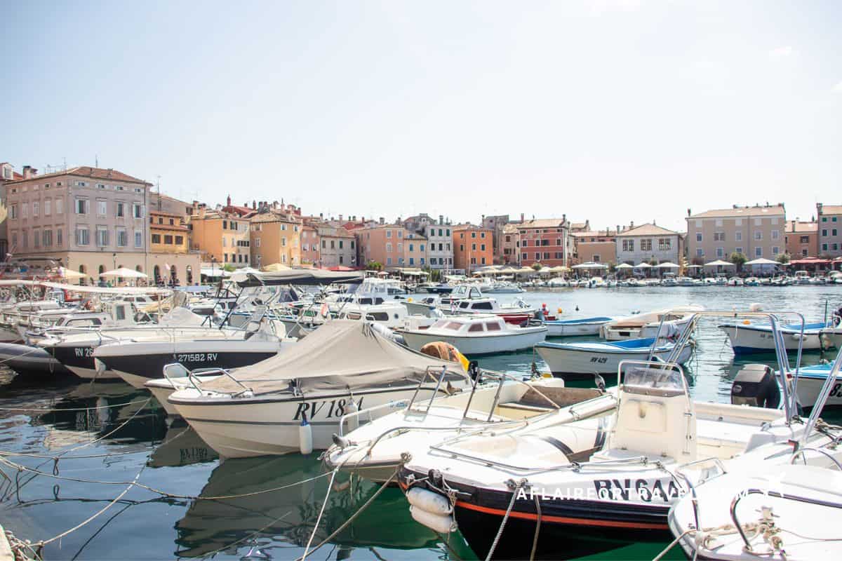 Small boats docked along the waterfront in Rovinj, Croatia, with pastel-colored buildings and cafés lining the harbor in the background.