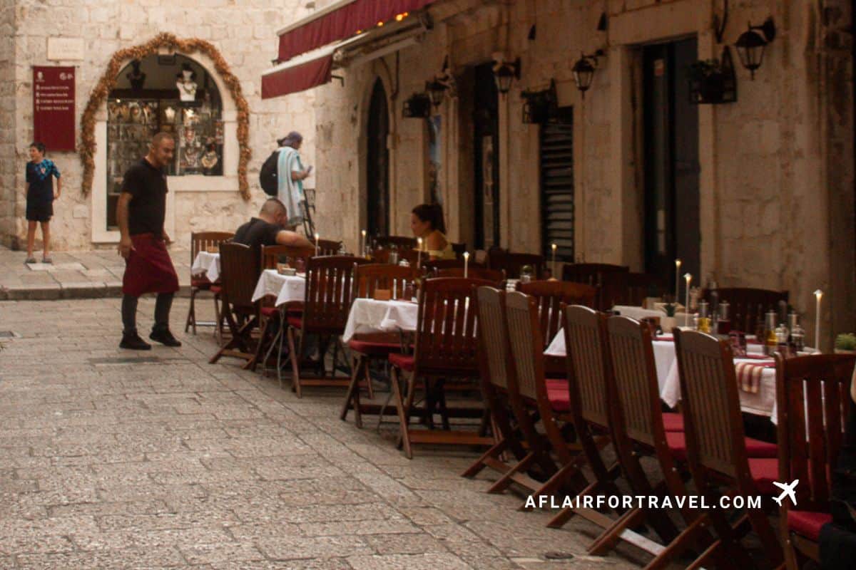 Outdoor restaurant seating along a quiet stone street in Dubrovnik Old Town with candlelit tables, historic buildings, and a server walking through the alley at dusk.