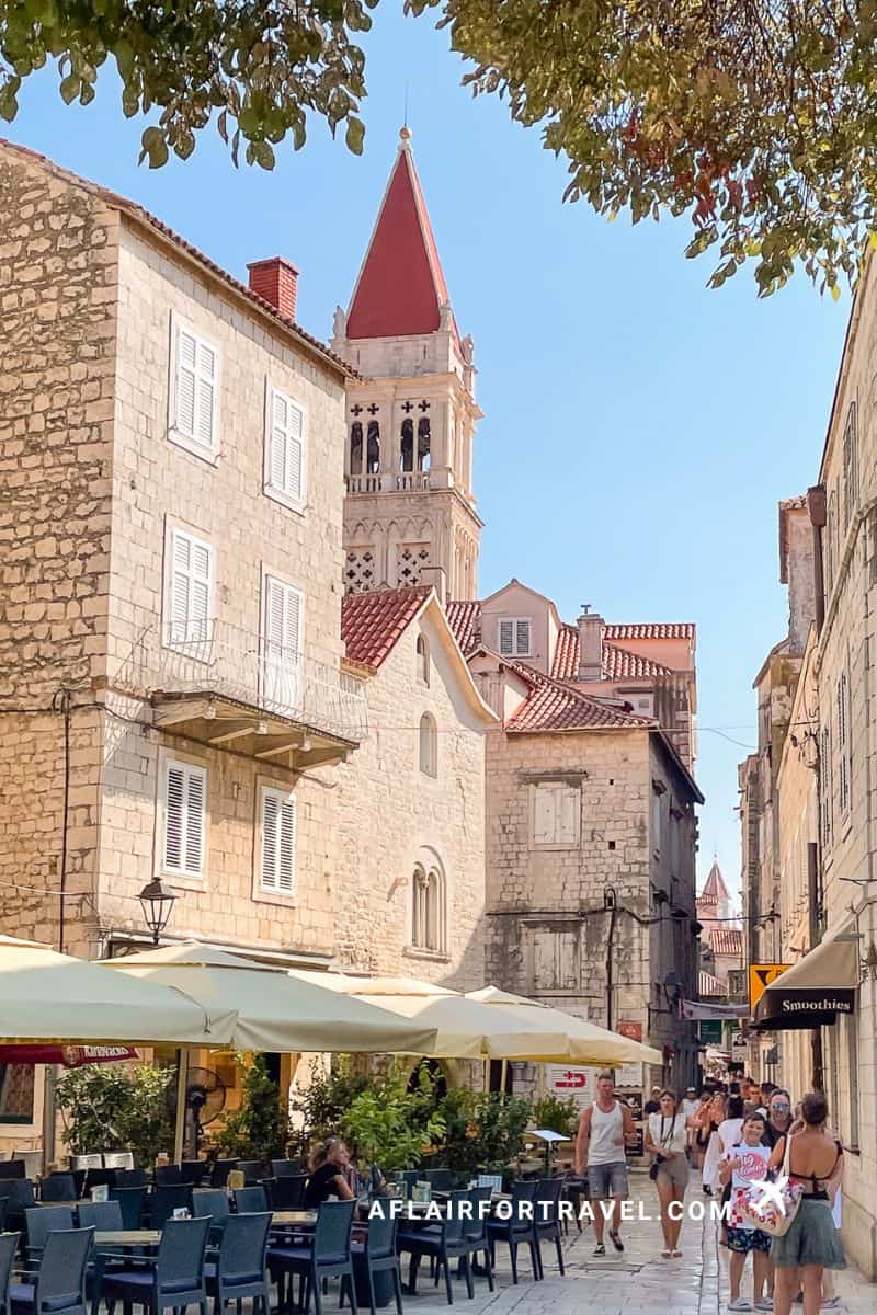 Tourists walking through the historic stone streets of Trogir's Old Town with a cathedral bell tower and outdoor café terrace, a UNESCO World Heritage site