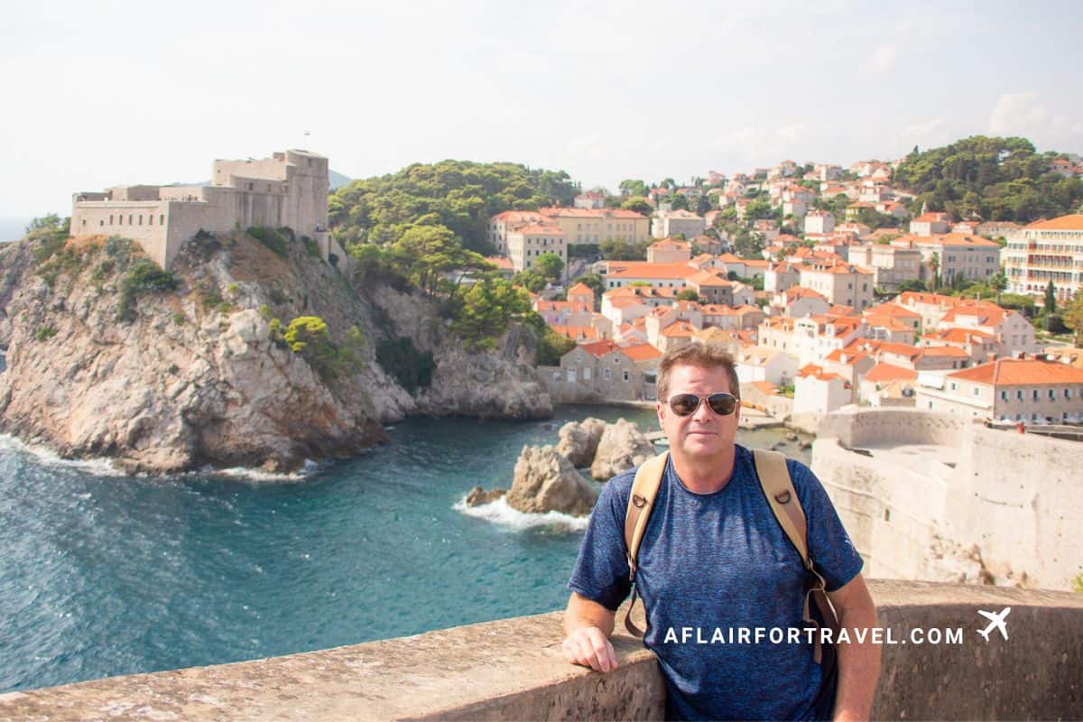 Man posing on Dubrovnik's ancient city walls with Fort Lovrijenac and the Old Town's terracotta rooftops in the background, a reminder to stay on designated paths and not climb on historic sites, one of the best examples of what not to do in Croatia