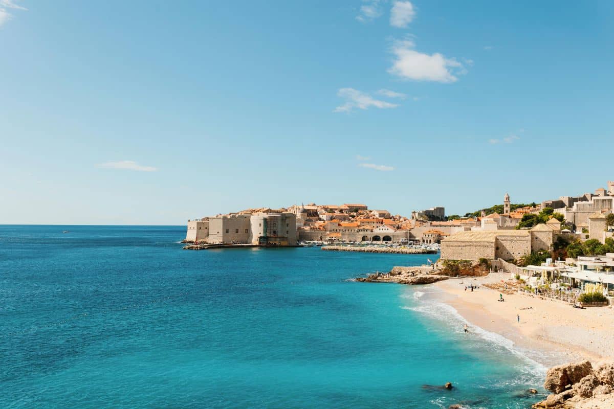 Panoramic view of Dubrovnik's iconic Old Town walls and sandy beach with vivid turquoise Adriatic waters.