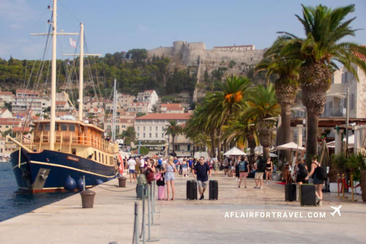 Busy waterfront promenade in Hvar Town with tourists, moored boats and a hilltop fortress, a popular Croatian destination